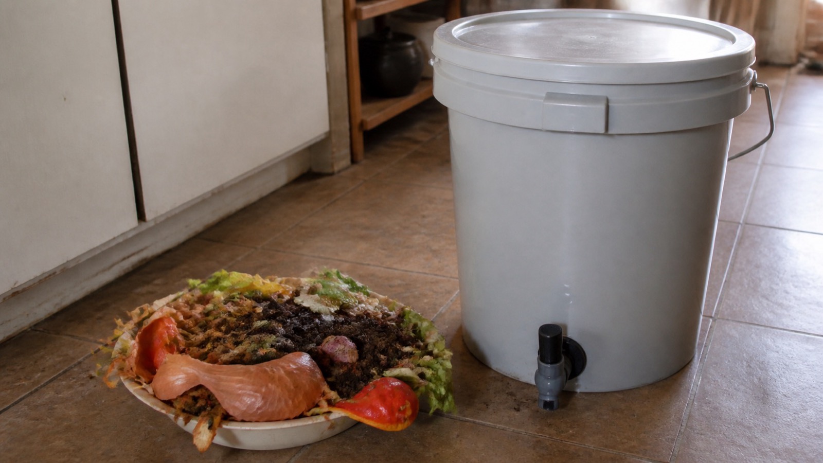 A sealed bokashi bucket with tap on a kitchen floor beside food scraps, bokashi