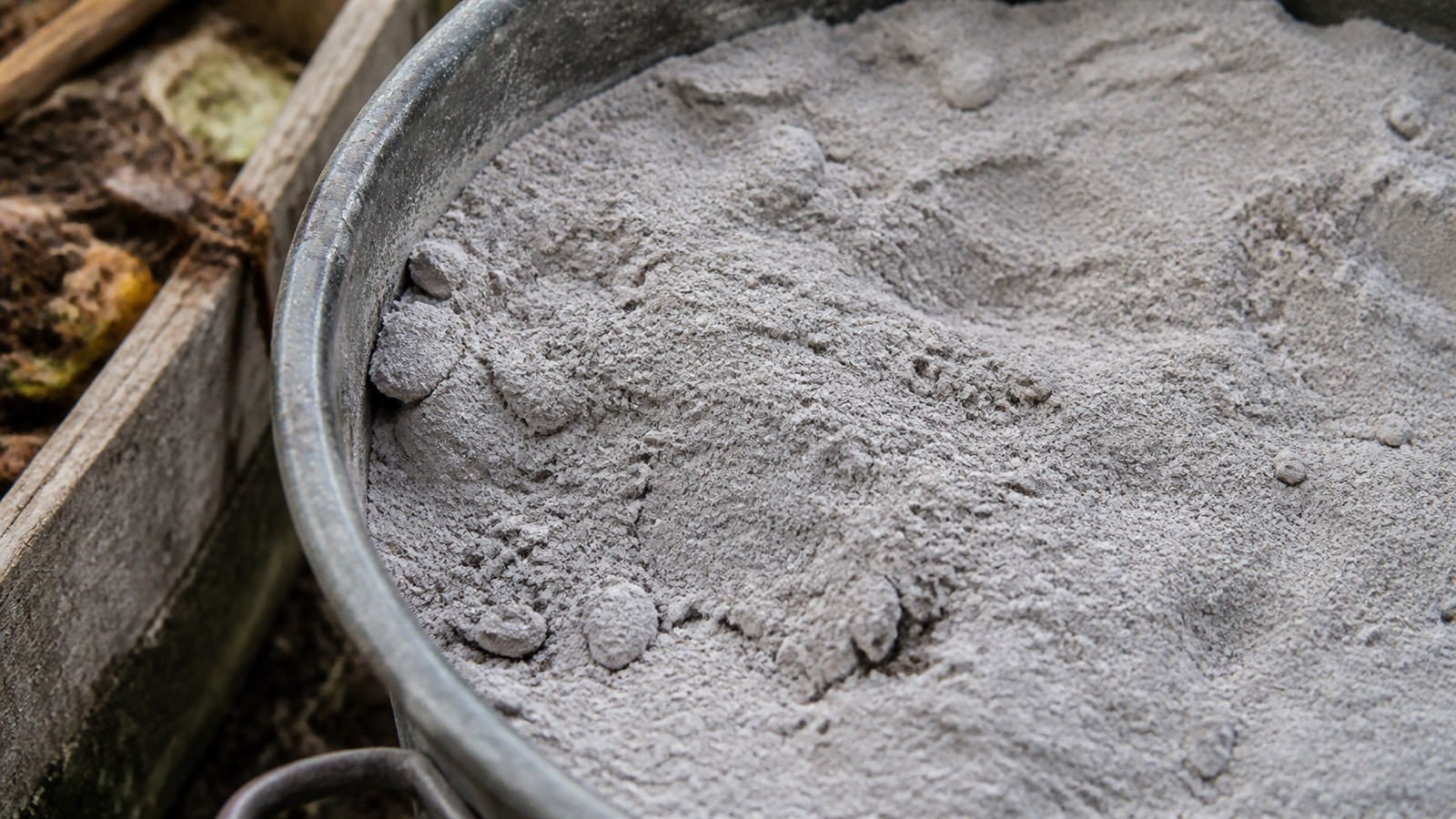 Extreme close-up filling the frame: pale grey wood ash in a metal bucket beside a compost bay, compost ash