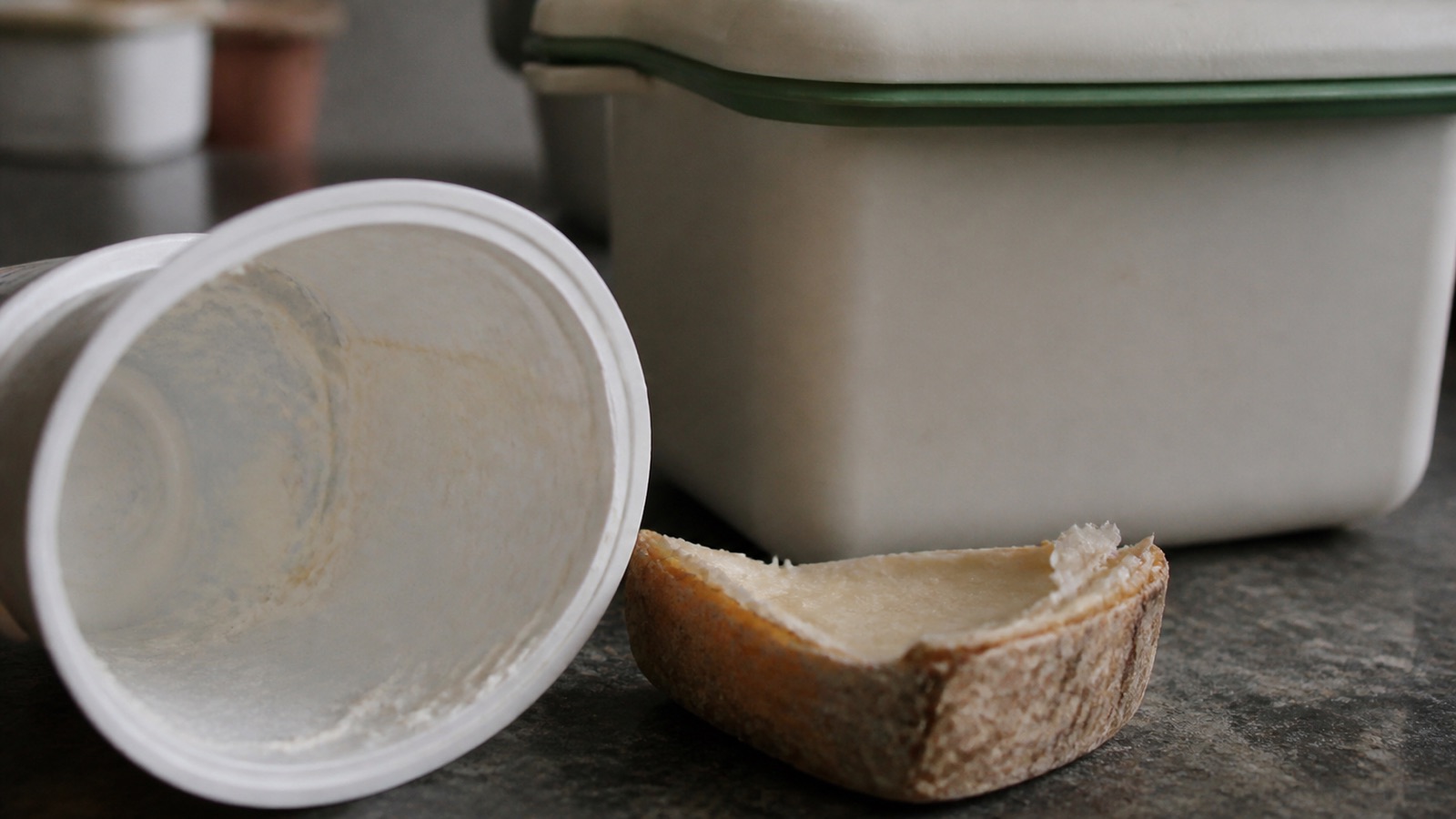 Extreme close-up filling the frame: an empty yoghurt pot and cheese rind on a kitchen counter beside a closed caddy, compost dairy